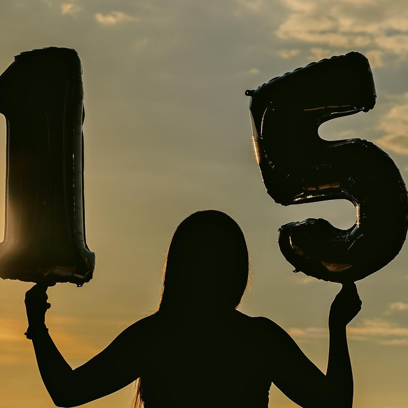 Silhouetted woman holding '15' balloons against a sunset backdrop, symbolizing celebration.